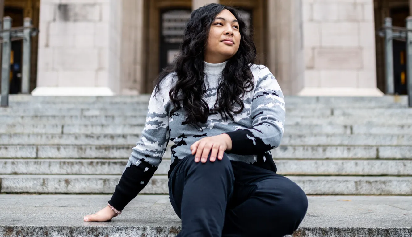 Headshot of Violet Monserate, a brown woman with long black hair, sitting on concrete stair steps in front of Suzallo Library